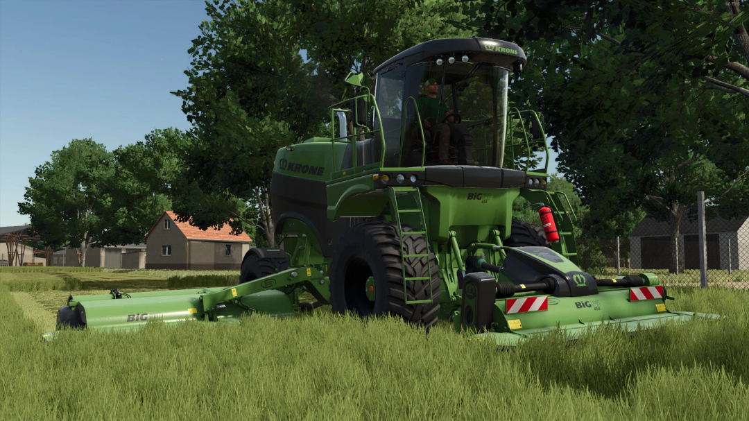 Farming Simulator 25 mod: Krone BigM v1.0.1.0 forage harvester in a field with green grass, trees, and buildings in the background.
