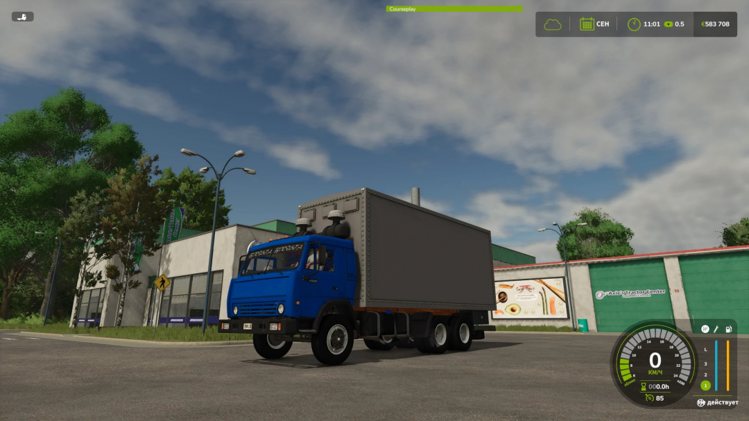 Farming Simulator 25 mod featuring the blue KamAZ 53212 truck with gray cargo container, parked outside a green building under a partly cloudy sky.