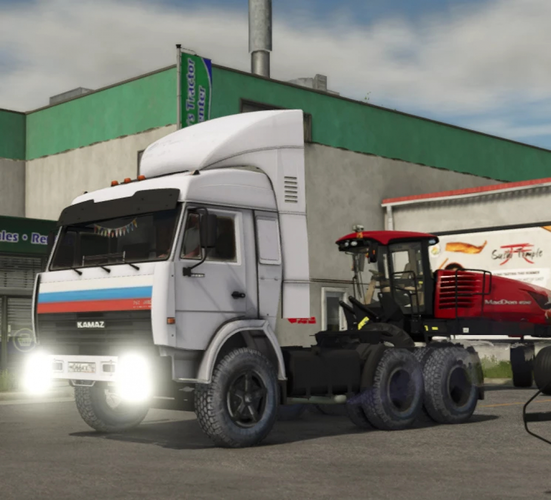 Farming Simulator 25 mods featuring the KamAZ 54115 truck mod, shown with a red tractor attached, parked in front of a green building.