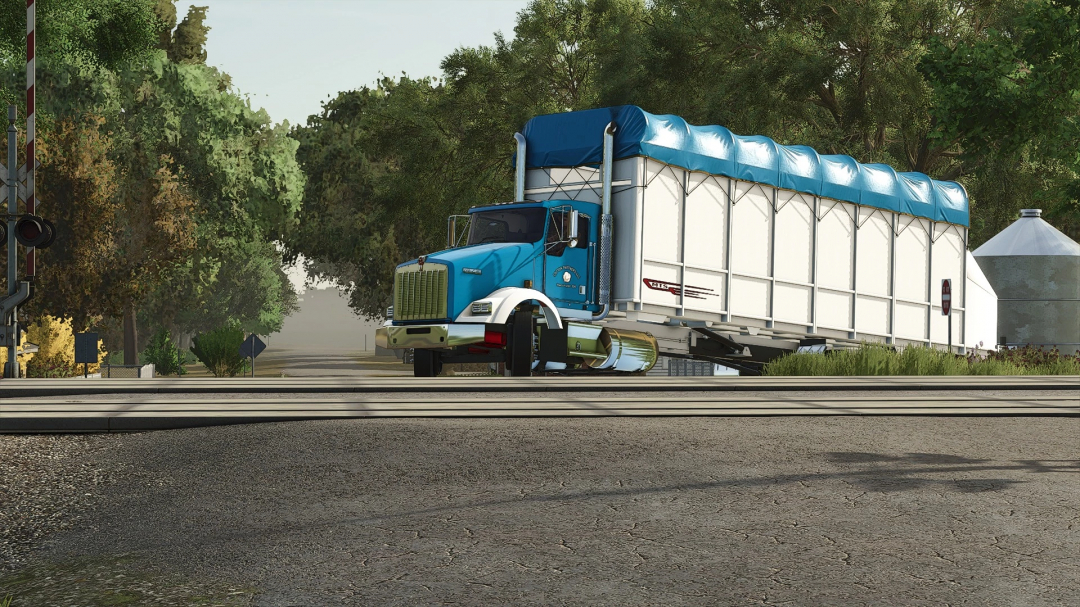 Farming Simulator 25 mod featuring a blue Kenworth KW900 Cotton Truck parked on a railway crossing with trees and farm structures in the background.